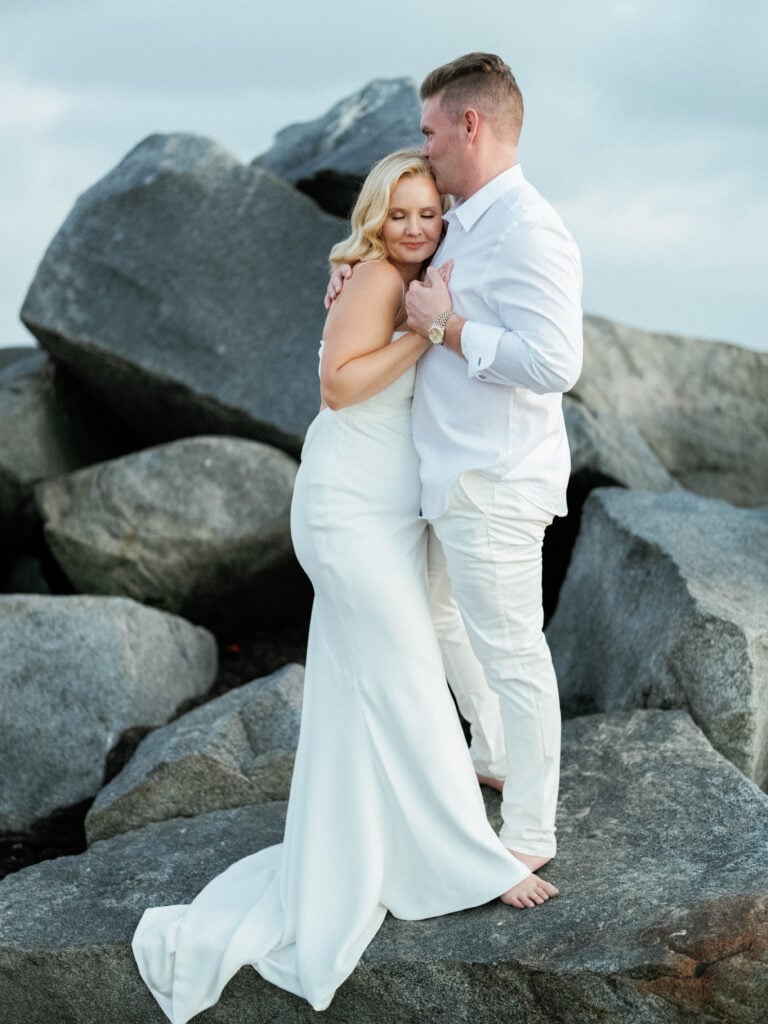 Bride in a strapless gown and groom in a white shirt embracing on large beach rocks, shot on film by a Palm Springs wedding photographer.