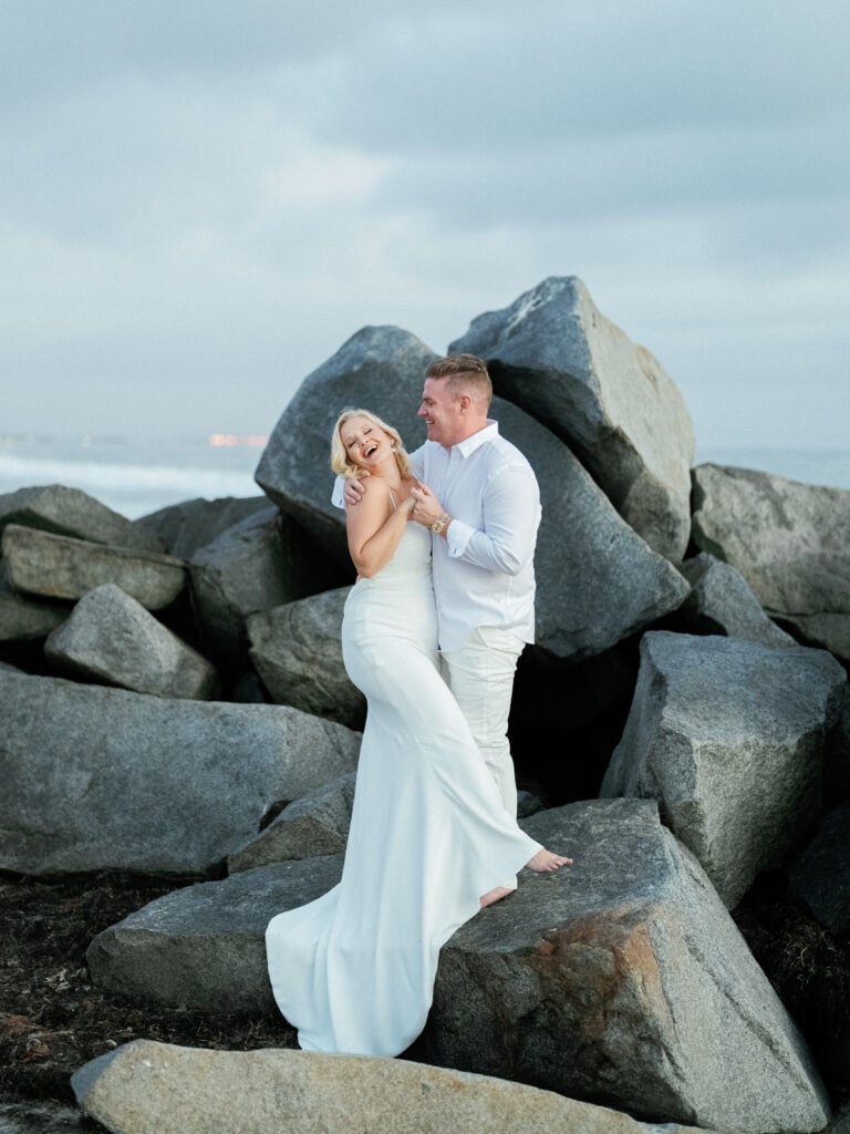 Bride and groom laughing while posing on large coastal boulders under a cloudy sky, captured on film by a Palm Springs wedding photographer.