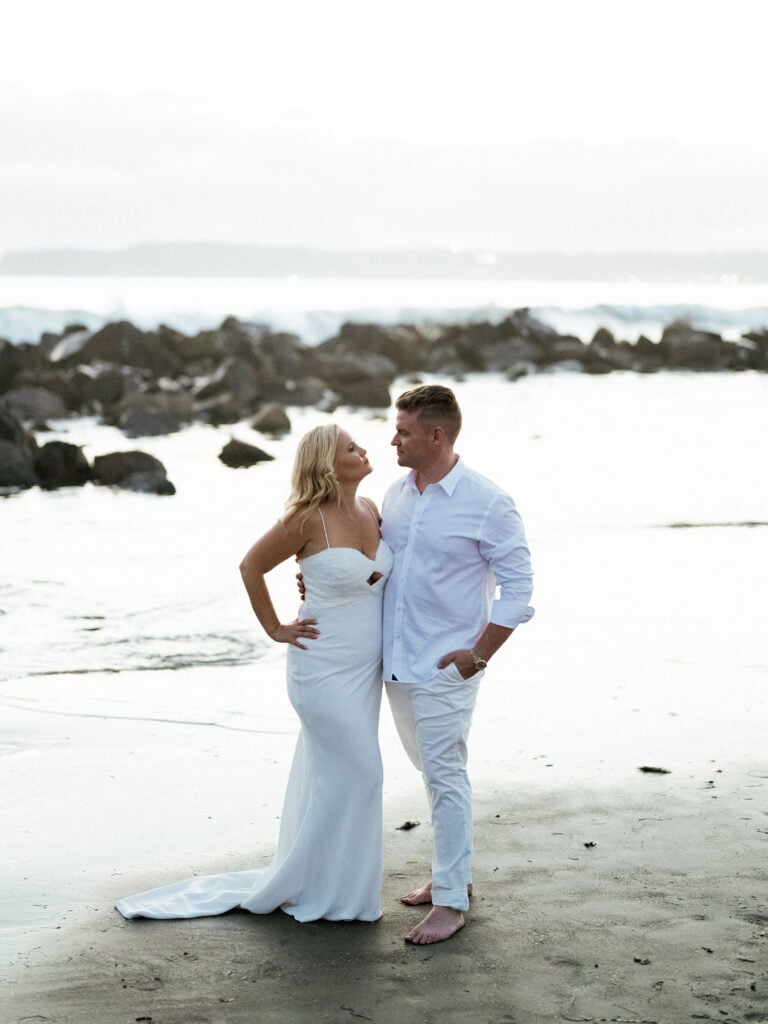 Bride and groom in all-white wedding attire standing on a wet sandy beach near rocks, shot on film by a Palm Springs wedding photographer.