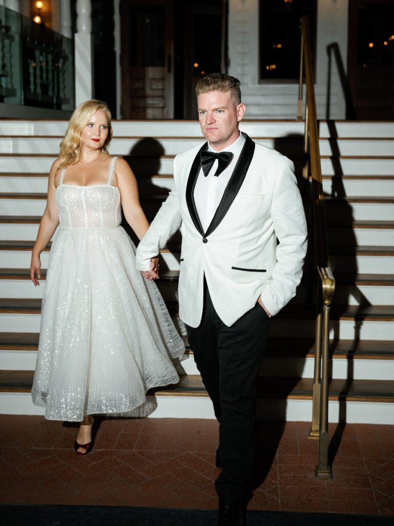 Bride in a midi sparkly dress and groom in a white tuxedo jacket walking down stairs, shot on film with flash by a Palm Springs wedding photographer.