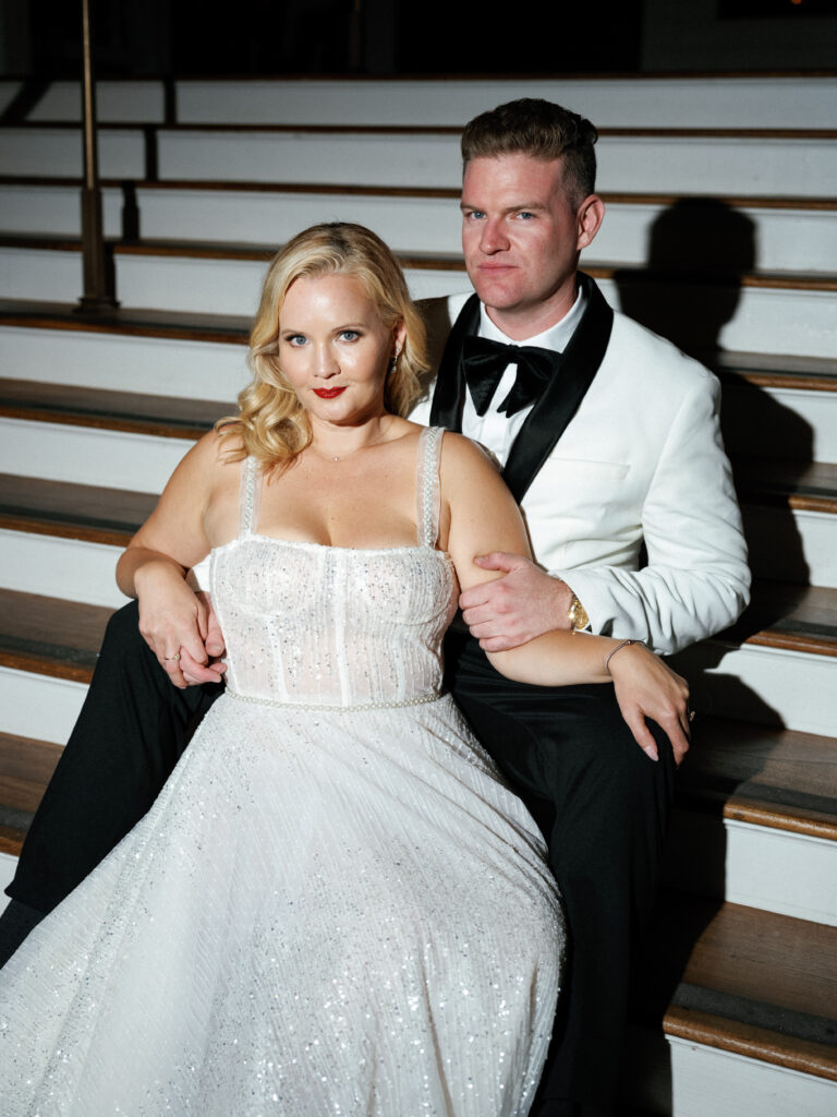 Elegant bride in a sparkly corset wedding dress and her groom in a white tuxedo jacket sitting on indoor stairs, showcasing a glamorous look for editorial wedding photography.