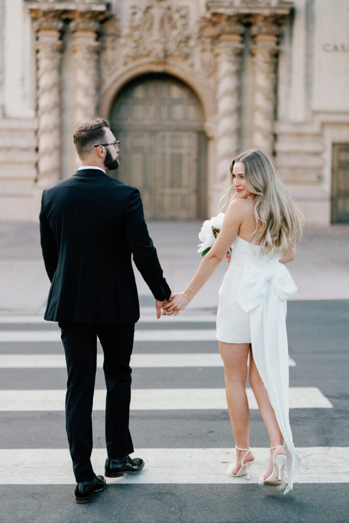 Elegant bride in a mini wedding dress with a long bow train holding hands with her groom while walking across a crosswalk, capturing authentic moments for editorial wedding photography.
