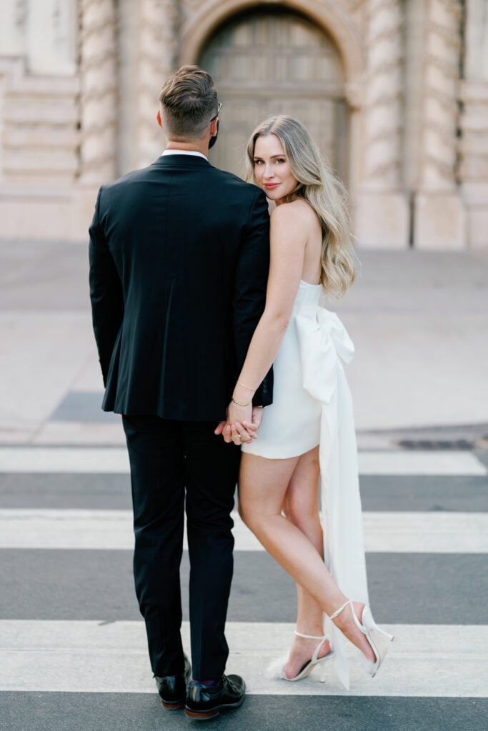 Candid bride looking over her shoulder in a short white wedding dress with a large statement bow back while crossing the street with her groom during an editorial wedding photography shoot.