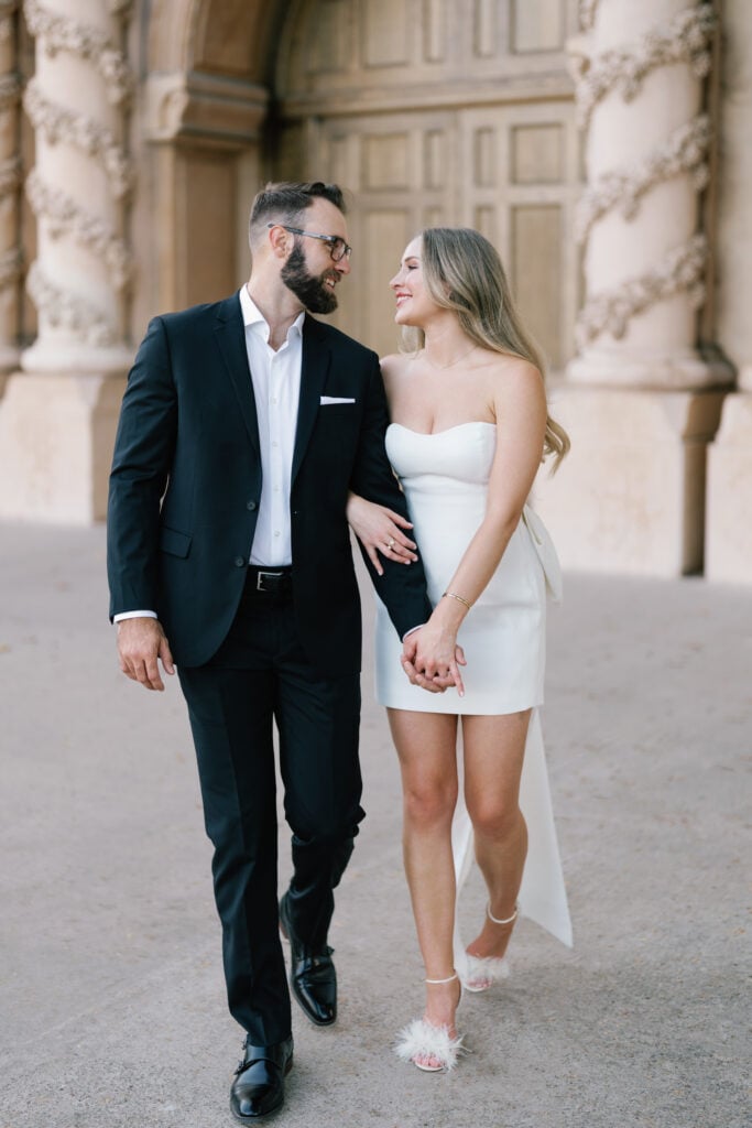 Elegant bride in a strapless mini wedding dress with a bow train walks hand-in-hand with her groom, capturing authentic moments for modern editorial wedding photography.