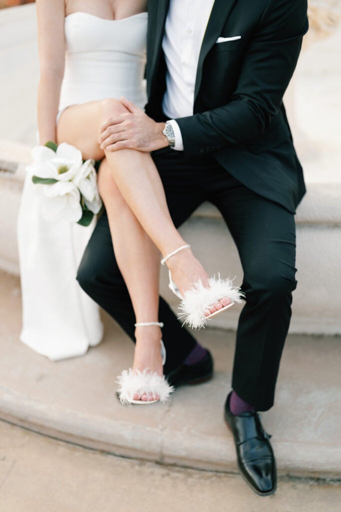 Close-up of a bride in chic white feather bridal sandals showing her engagement ring while leaning on her groom, highlighting high-end details in editorial wedding photography.