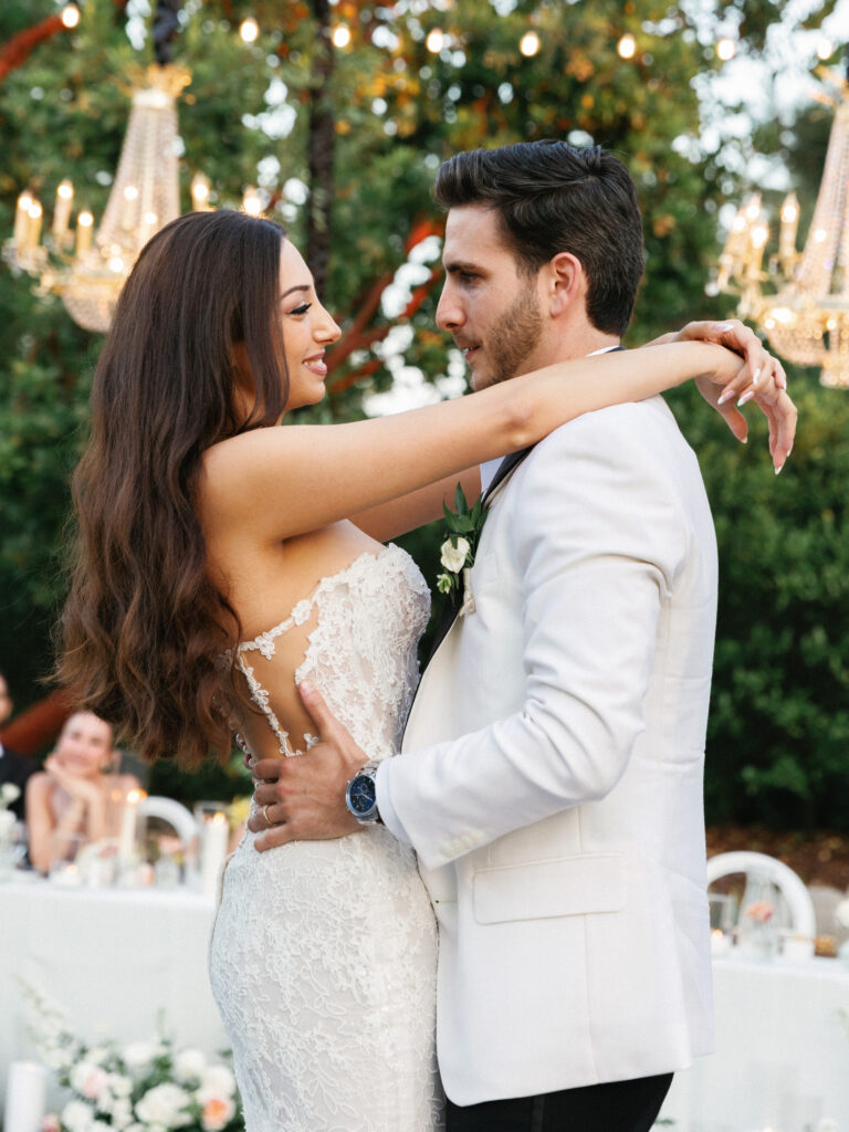Romantic couple hugging while enjoying their wedding first dance under warm outdoor reception lights