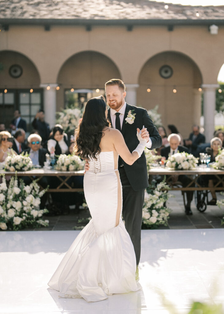 Bride in a mermaid dress and groom smiling during their wedding first dance on a white outdoor dance floor