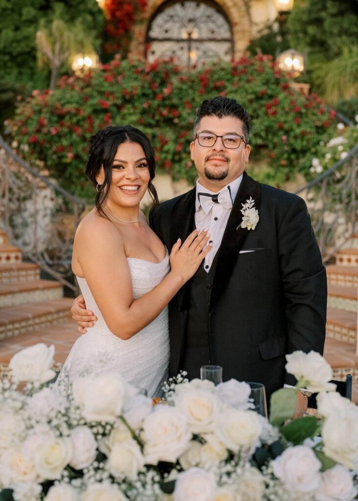 Bride and groom smiling at their sweetheart table filled with white roses during wedding reception toasts
