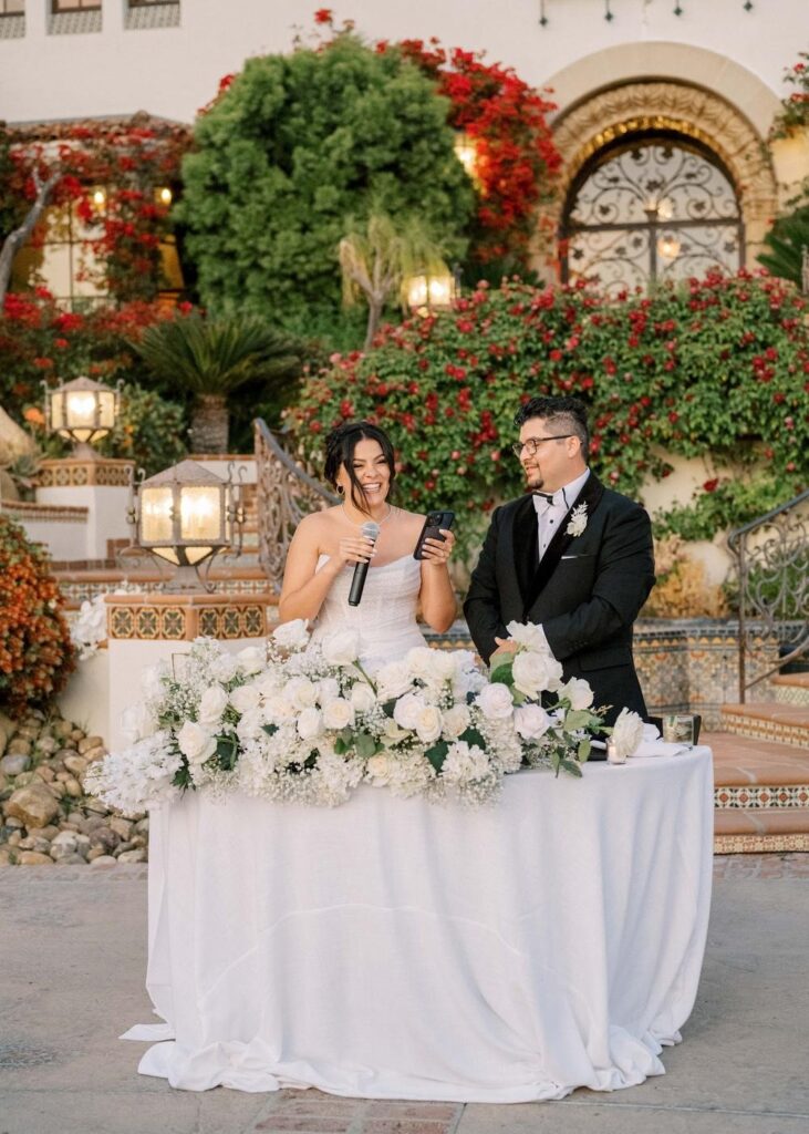 Happy newlyweds standing at their floral sweetheart table for wedding reception toasts and welcome speech