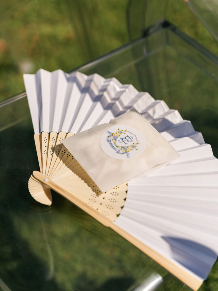 Paper hand fan and custom program favor placed on a clear chair as part of smart texas summer wedding tips for guest comfort
