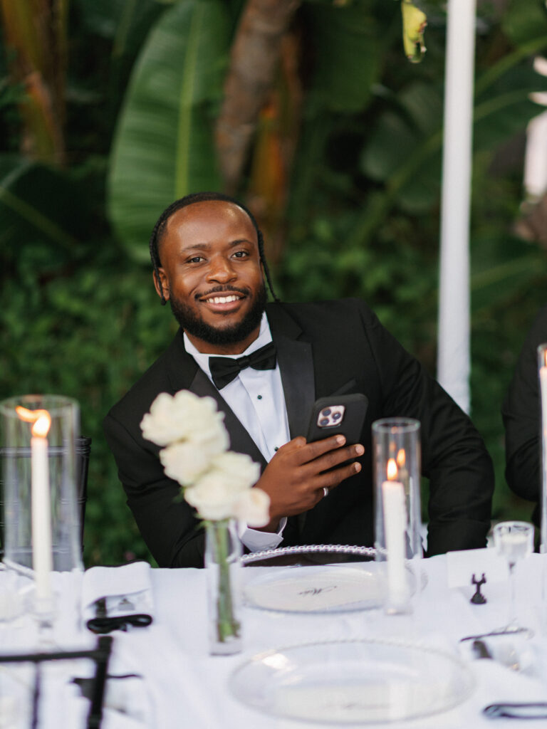 Smiling man in a black tuxedo holding a phone at the dinner table during wedding reception toasts
