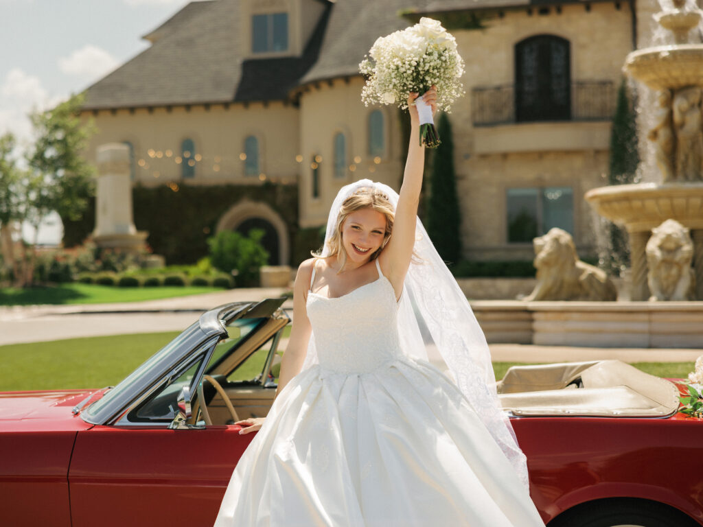 Bride celebrating in a classic vintage getaway car with a white rose bouquet outside a luxury estate venue