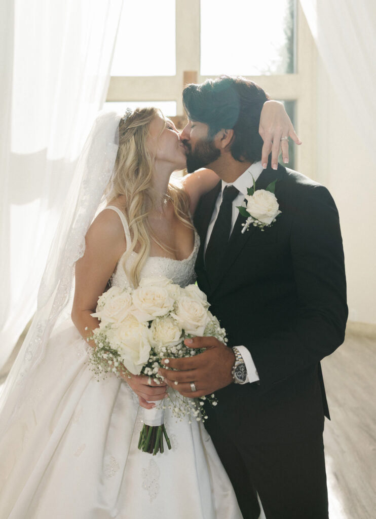 Indoor wedding photo session away from heat featuring a romantic bride and groom kissing in front of bright windows