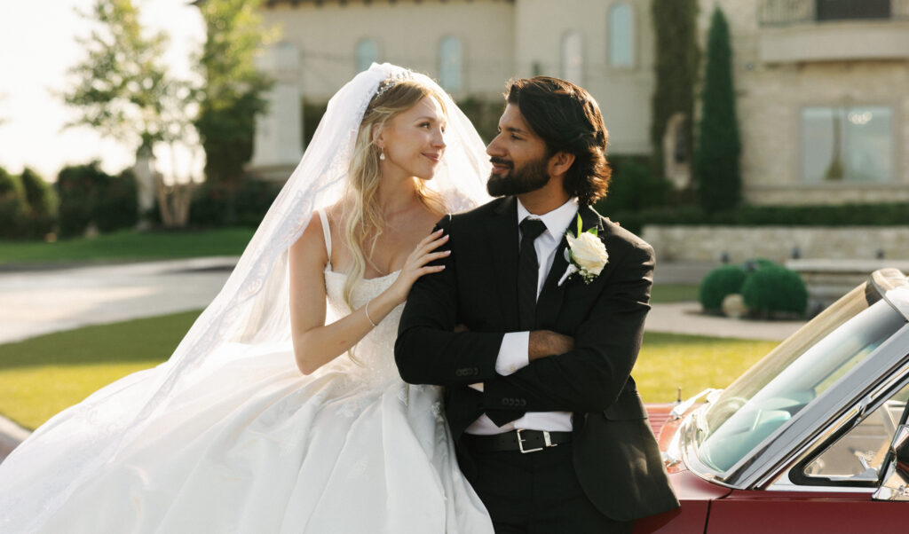 Newlyweds looking at each other while posing by a classic convertible during golden hour portrait session
