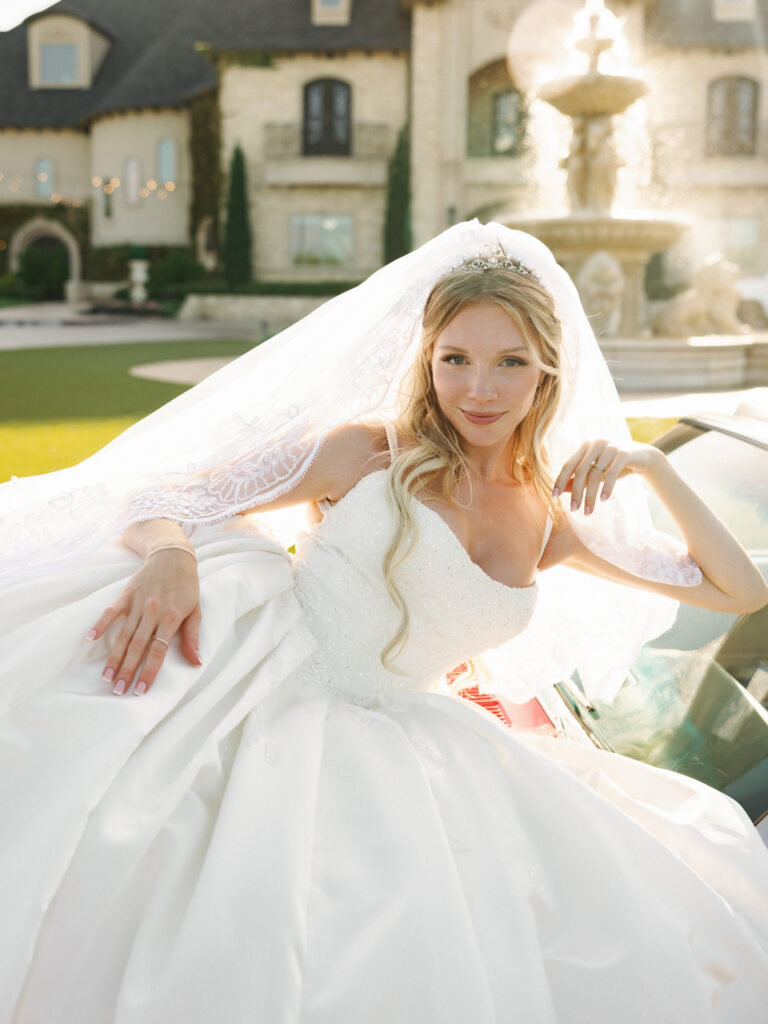 Bride wearing a luxury ballgown and an elegant lace veil while sitting in a classic getaway car