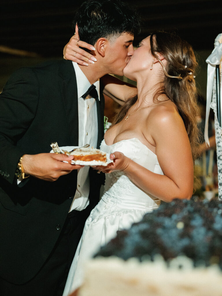 Newlyweds kissing passionately while holding a plate with a slice of wedding cake