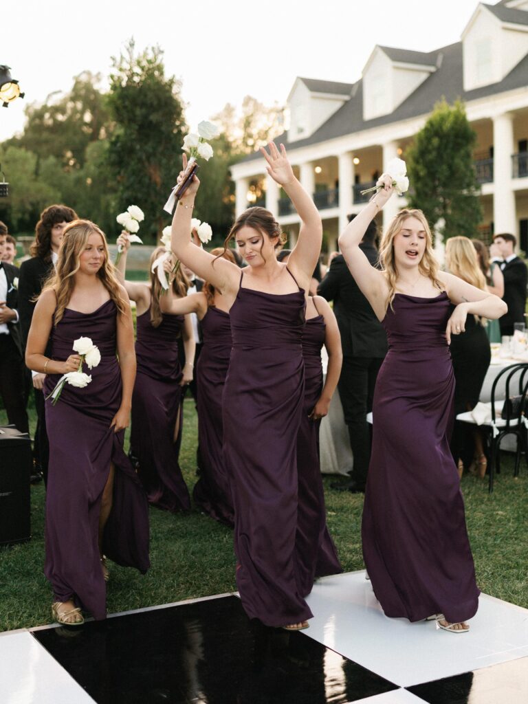 Bridesmaids in plum purple satin dresses dancing and celebrating on an outdoor wedding dance floor