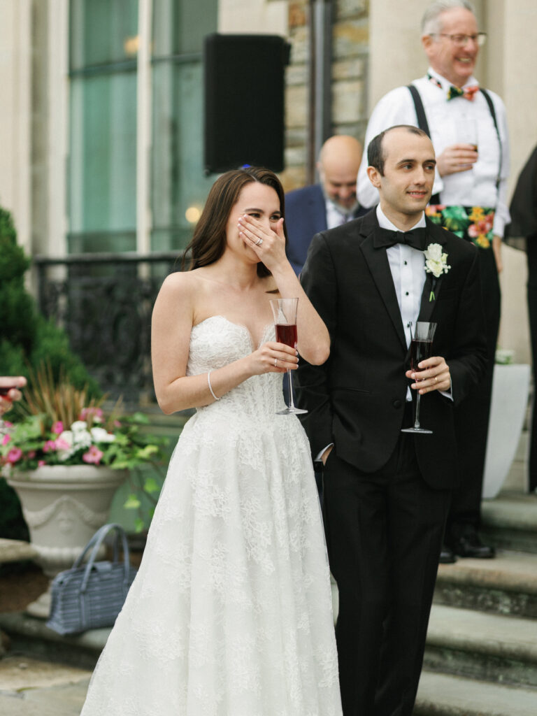 Bride laughing and covering her face while listening to wedding reception toasts with her groom outdoors