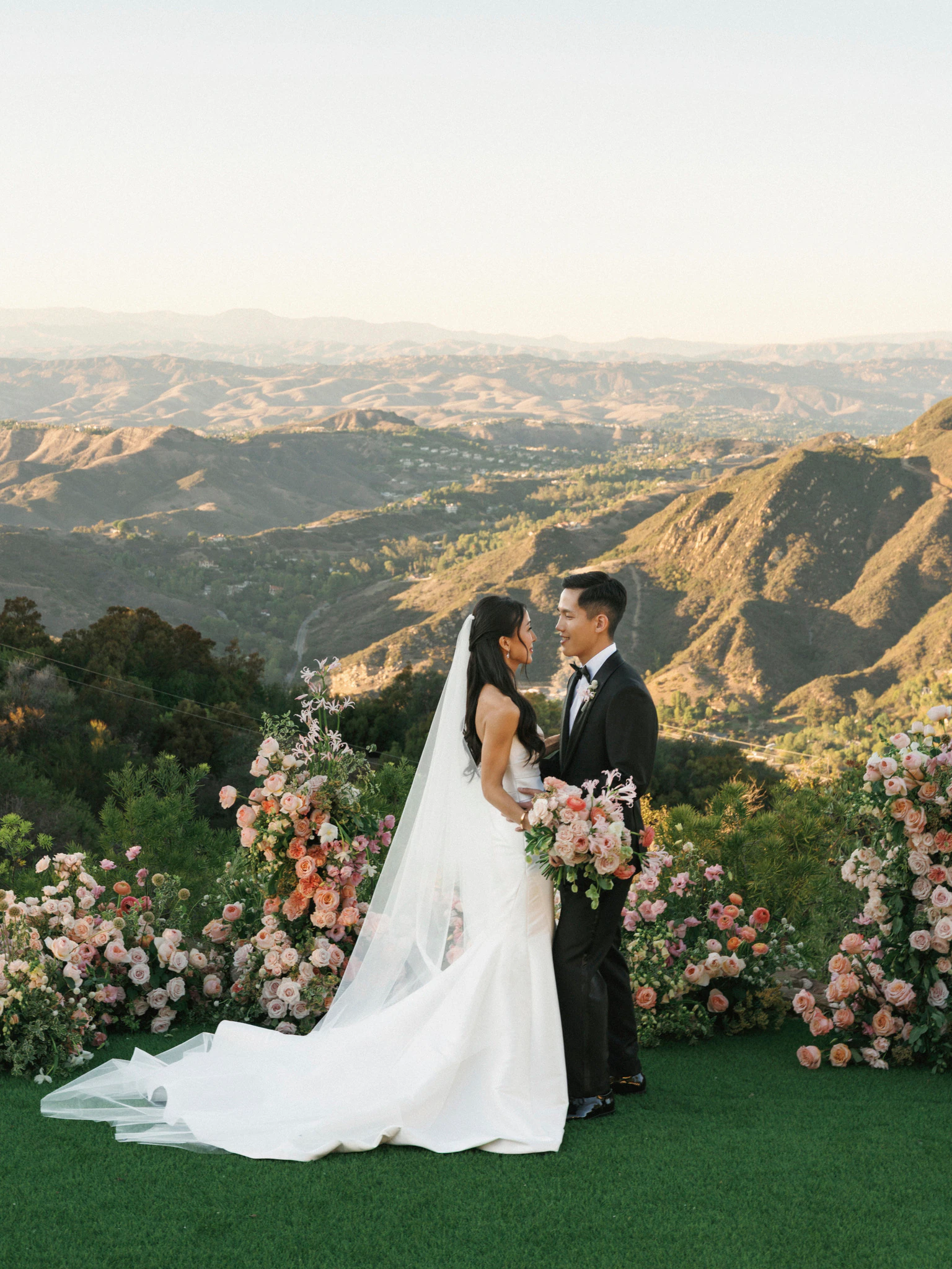Newlywed couple standing amid floral arrangements with a sunlit mountain landscape