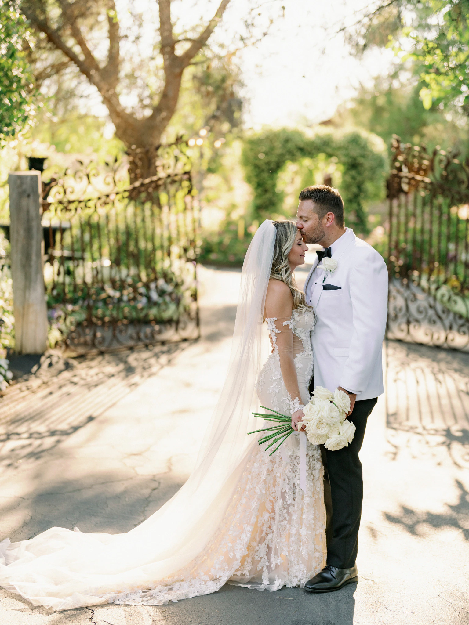 Bride and groom standing beneath sunlit trees beside an iron gate